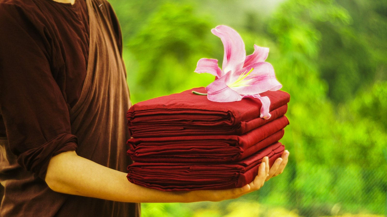 A person in robes holding a stack of folded new robes with a pink lily resting on top